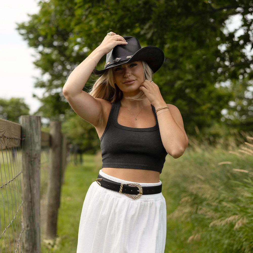 Woman wearing a black cowboy hat, black top, and white skirt in a grassy outdoor setting.