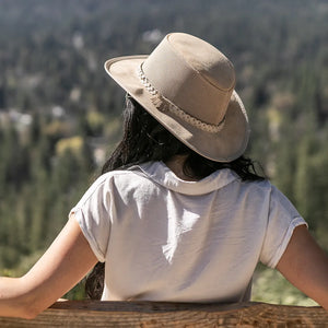 Person wearing a beige hat and white shirt, sitting on a wooden bench with a forest view.