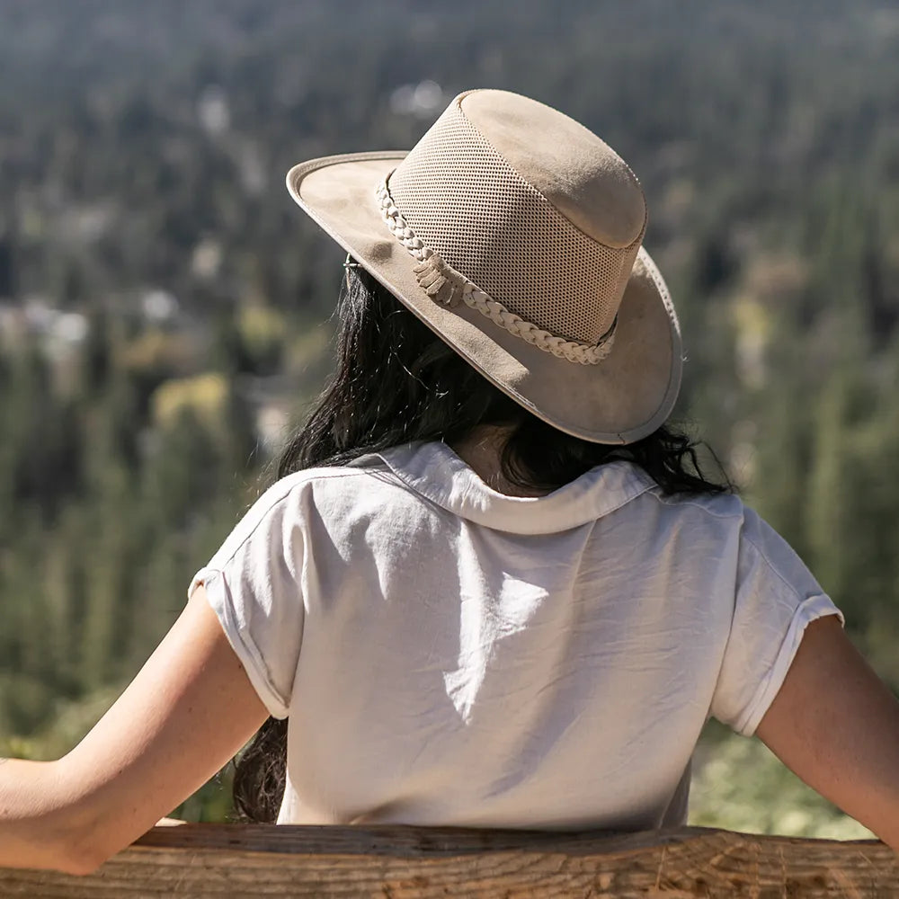 Person wearing a beige hat and white shirt, sitting on a wooden bench with a forest view.