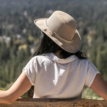 Person wearing a beige hat and white shirt, sitting on a wooden bench with a forest view.
