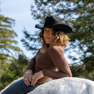 Woman wearing a black cowboy hat and brown top sitting on a horse with trees in the background