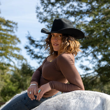 Woman wearing a black cowboy hat and brown top sitting on a horse with trees in the background