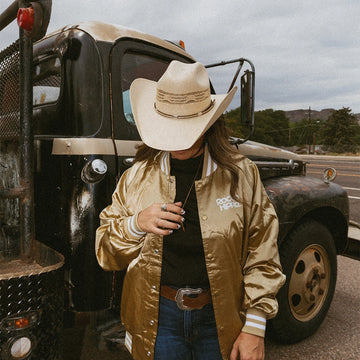Person wearing a cowboy hat and gold jacket standing next to an old truck on a road.