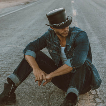 Man sitting on a road wearing a denim jacket, black top hat, and boots.