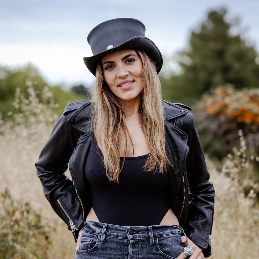 Woman wearing a black hat, leather jacket, and jeans standing in a natural setting.