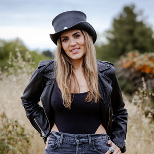 Woman wearing a black hat, leather jacket, and jeans standing in a natural setting.