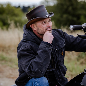 Man wearing a cowboy hat and dark jacket sitting on a motorcycle in a natural setting.