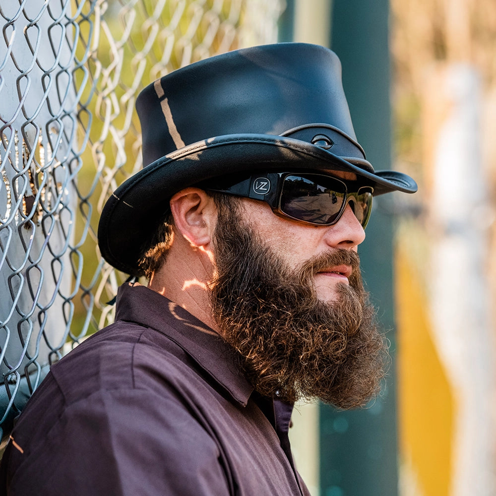 Man wearing a black hat and sunglasses with a beard, standing against a chain-link fence.