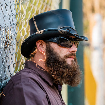 Man wearing a black hat and sunglasses with a beard, standing against a chain-link fence.