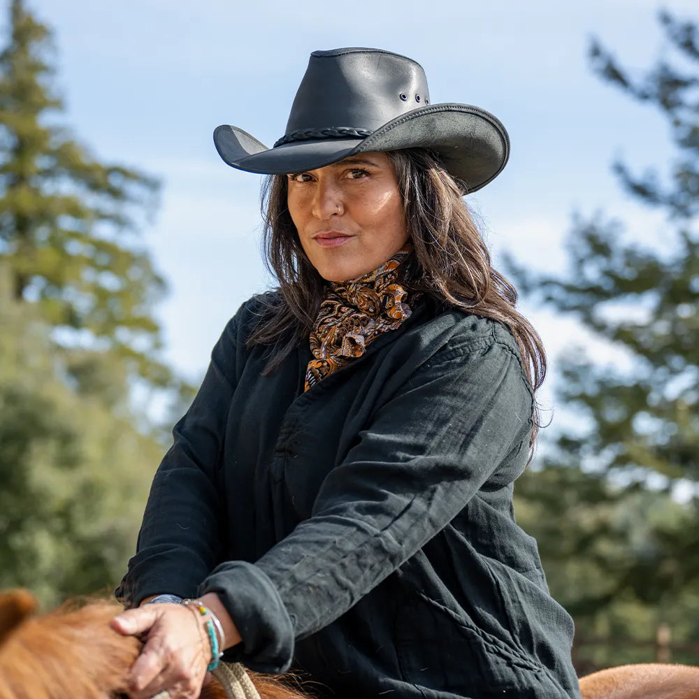 Woman in a cowboy hat and scarf sitting on a horse with trees in the background