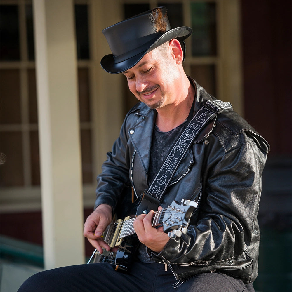 Man playing guitar wearing a black leather jacket and top hat outdoors.