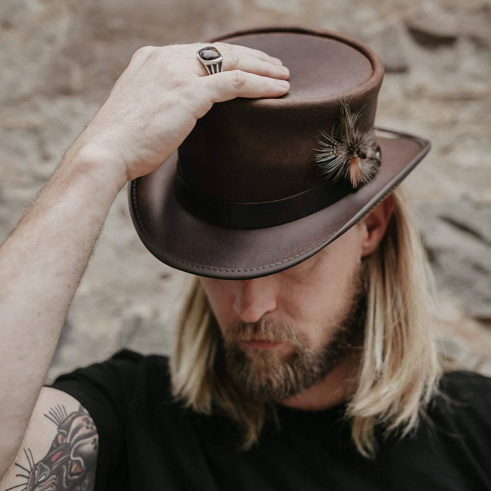 Man with tattoos adjusting a brown leather hat with a feather against a blurred natural background