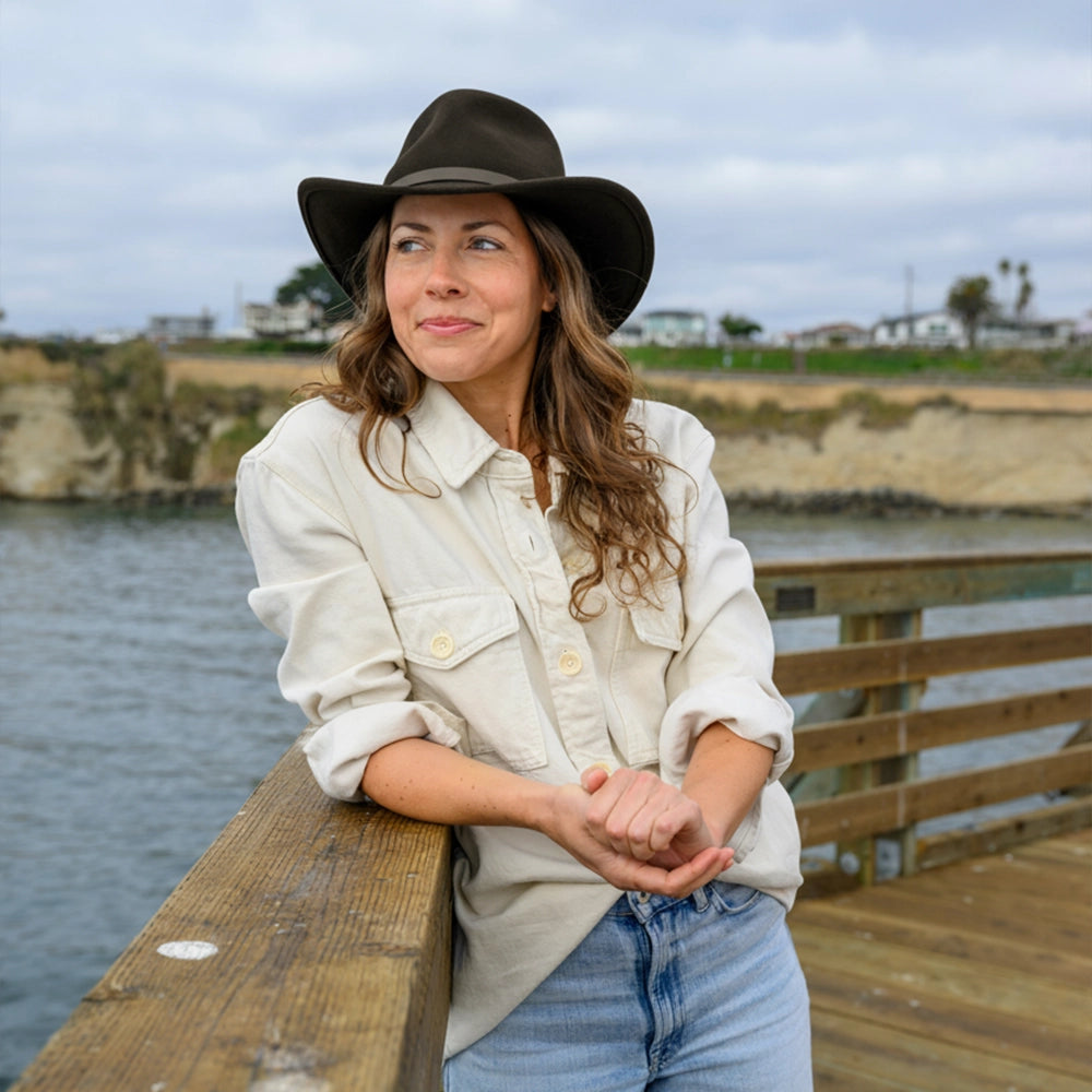 Woman wearing a black hat and light-colored shirt standing on a wooden dock by a body of water.