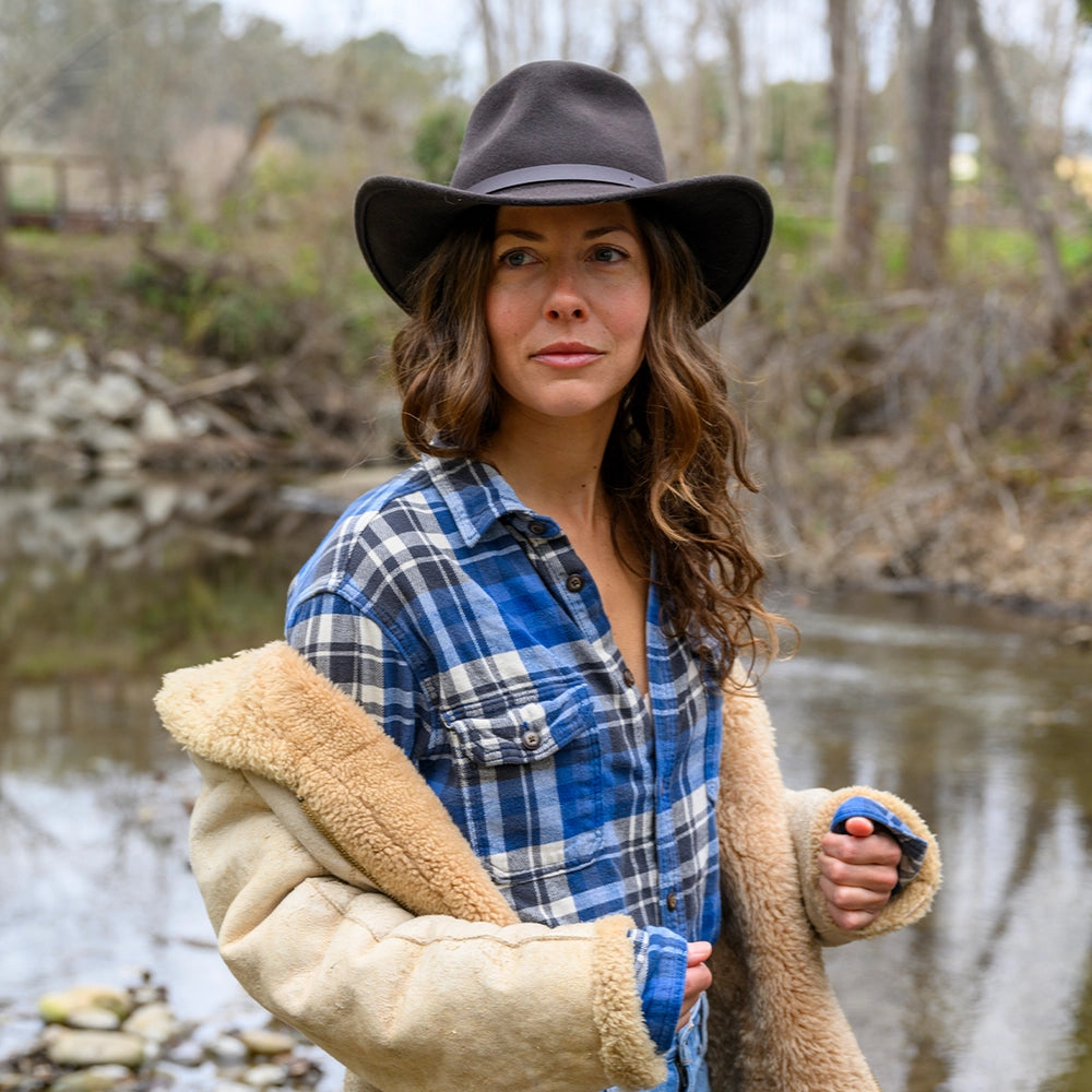 Woman wearing a plaid shirt, beige jacket, and dark hat standing by a body of water.