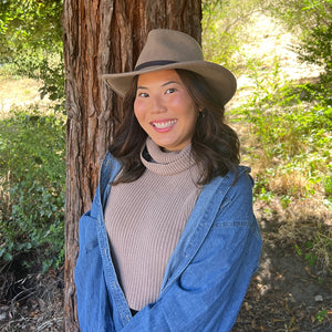 Woman wearing a beige hat and blue jacket standing next to a tree in a natural setting