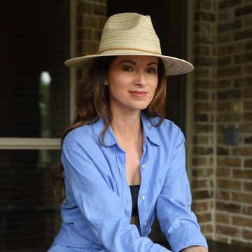 Woman wearing a straw hat and blue shirt sitting against a brick wall.