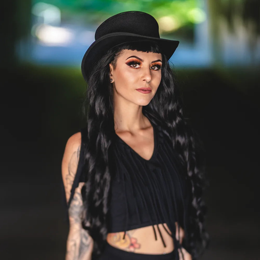 Woman wearing a black hat and black top with tattoos, standing outdoors.