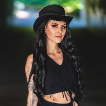 Woman wearing a black hat and black top with tattoos, standing outdoors.