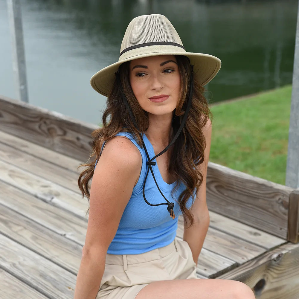 Woman wearing a blue top and beige hat sitting on a wooden dock by a body of water.