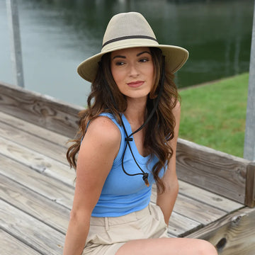 Woman wearing a blue top and beige hat sitting on a wooden dock by a body of water.