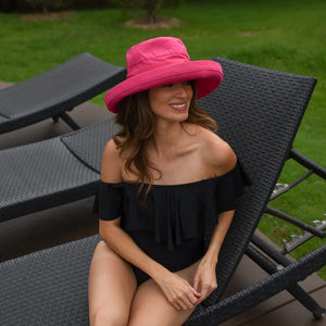 Woman wearing a black swimsuit and pink hat sitting on a lounge chair outdoors.