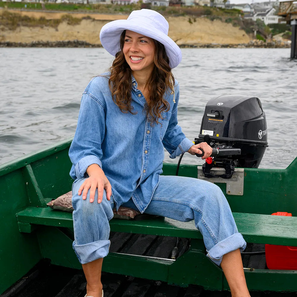Woman sitting on a boat by the water, wearing a blue denim outfit and white sun hat.