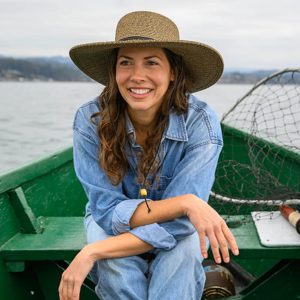 Woman wearing a wide-brimmed hat and denim jacket sitting in a green boat on water.
