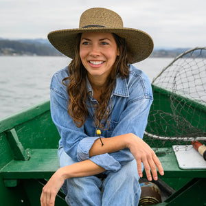 Woman wearing a wide-brimmed hat and denim jacket sitting in a green boat on water.