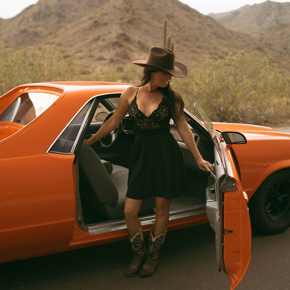 Woman in a black dress and cowboy hat getting out of an orange car in a desert setting