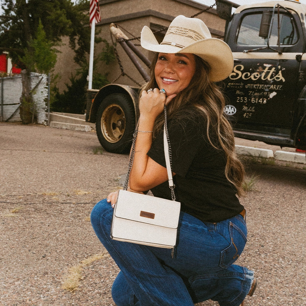 Woman in cowboy hat and jeans holding a white purse, with a truck and American flag in the background.
