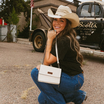 Woman in cowboy hat and jeans holding a white purse, with a truck and American flag in the background.