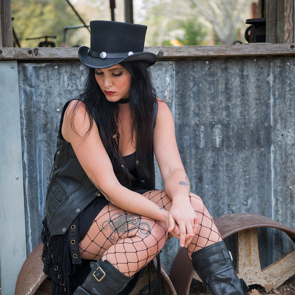 Woman in black leather outfit and top hat sitting against a rustic metal wall.
