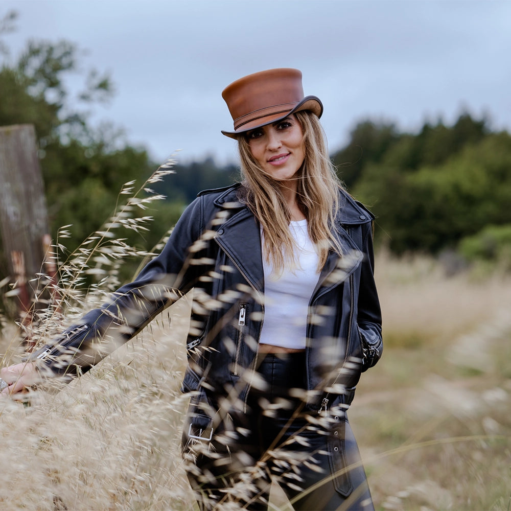 Woman in a leather jacket and hat standing in a field with tall grass.