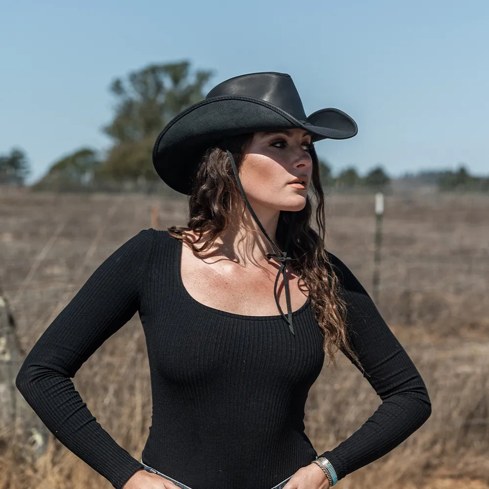 Woman wearing a black cowboy hat and black long-sleeve top standing in an open field.