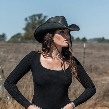 Woman wearing a black cowboy hat and black long-sleeve top standing in an open field.