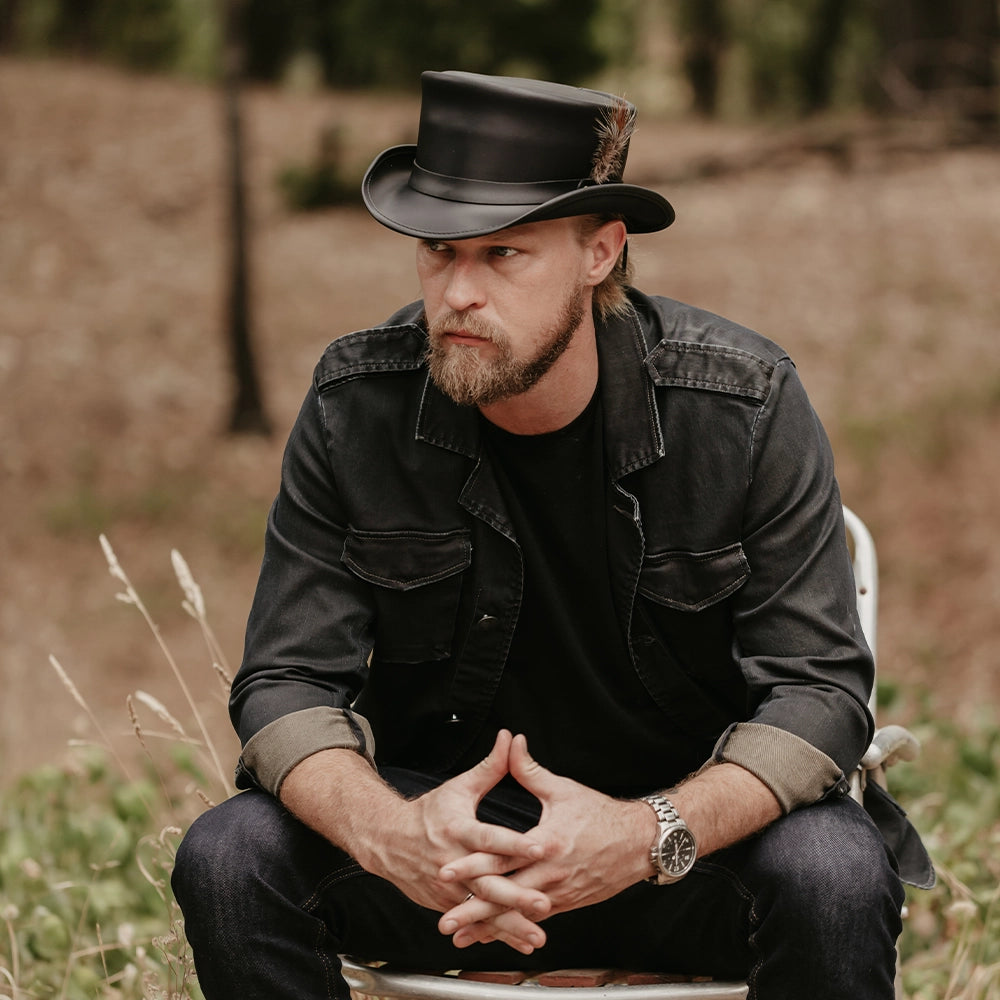 Man wearing a black hat and jacket sitting outdoors with a blurred natural background