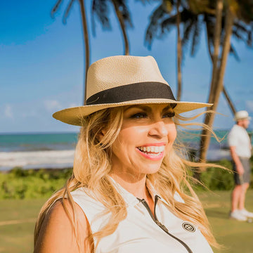 Woman wearing a straw hat with a black band, standing outdoors with palm trees and ocean in the background.