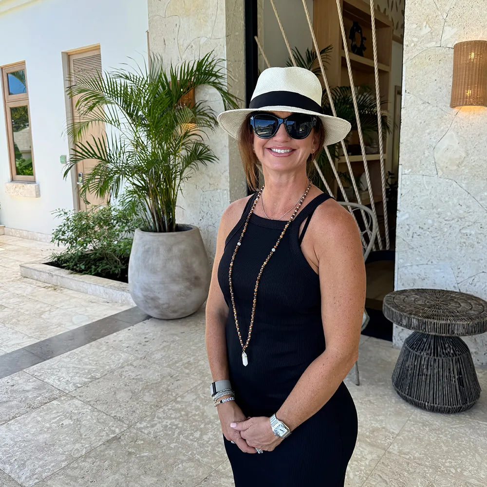 Woman in a black dress and hat with sunglasses standing outdoors.