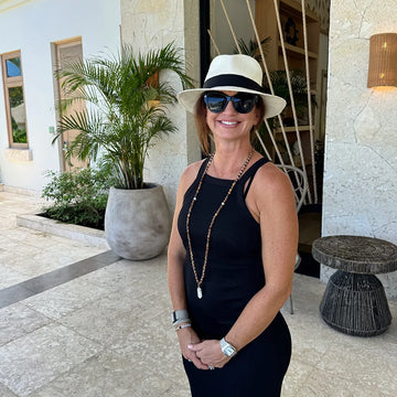 Woman in a black dress and hat with sunglasses standing outdoors.