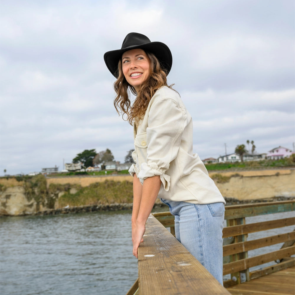 Woman in a black hat and light jacket standing on a wooden pier by a body of water.