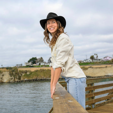 Woman in a black hat and light jacket standing on a wooden pier by a body of water.