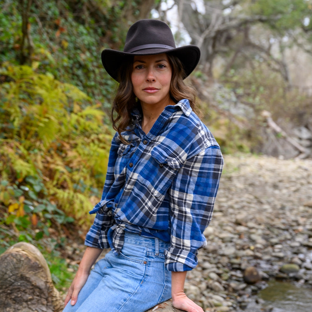Woman wearing a blue plaid shirt and black hat sitting on a rock in a natural setting.