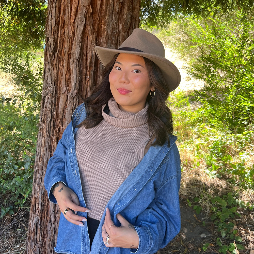 Woman wearing a beige hat and blue jacket standing next to a tree in a forest.