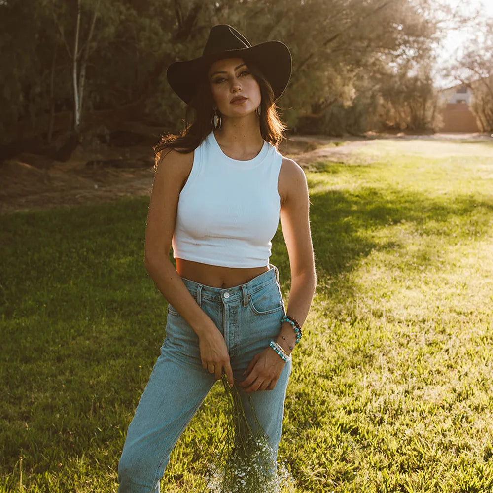 Woman in a white tank top and black hat standing in a grassy field with trees in the background.
