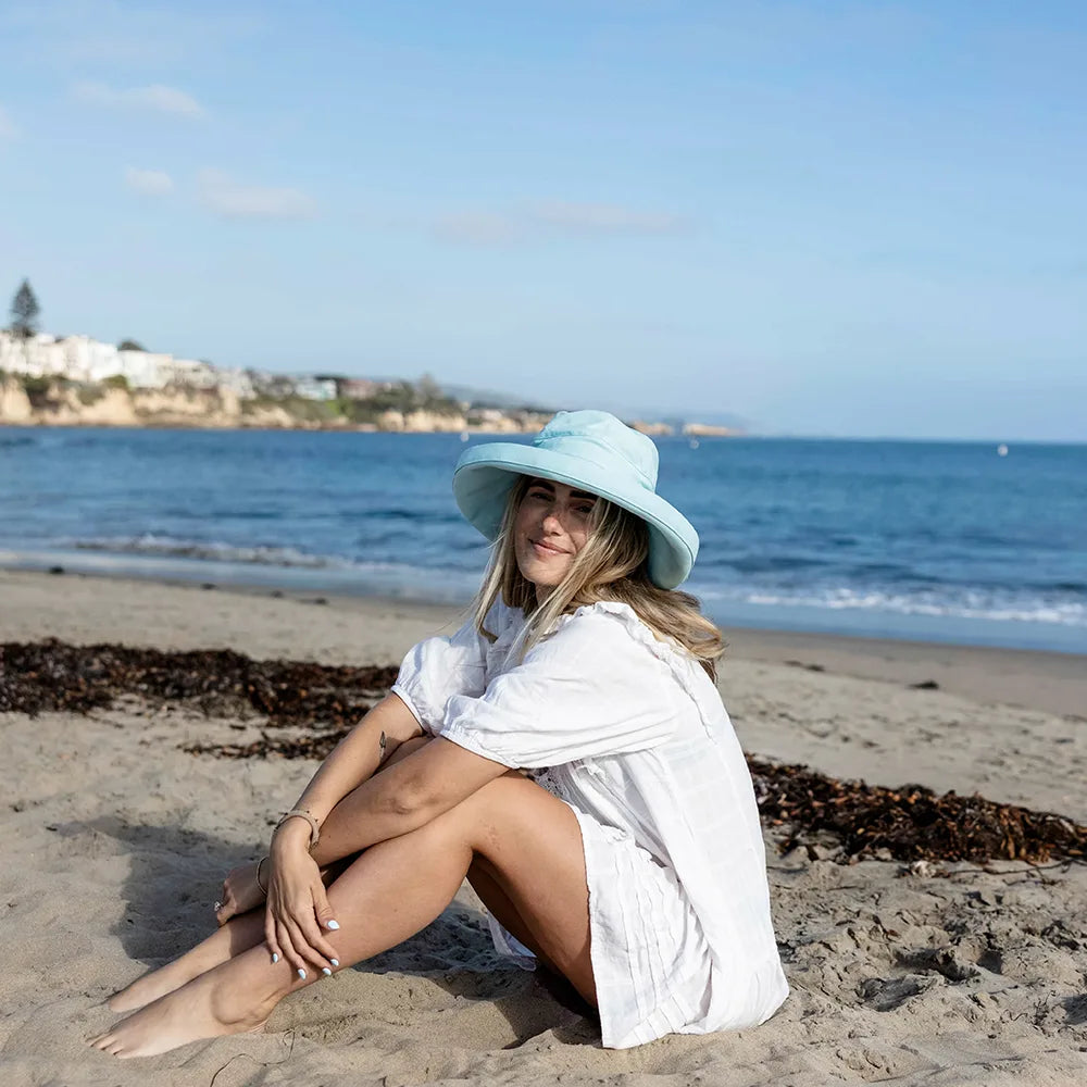 Woman sitting on a beach wearing a light blue hat and white cover-up.
