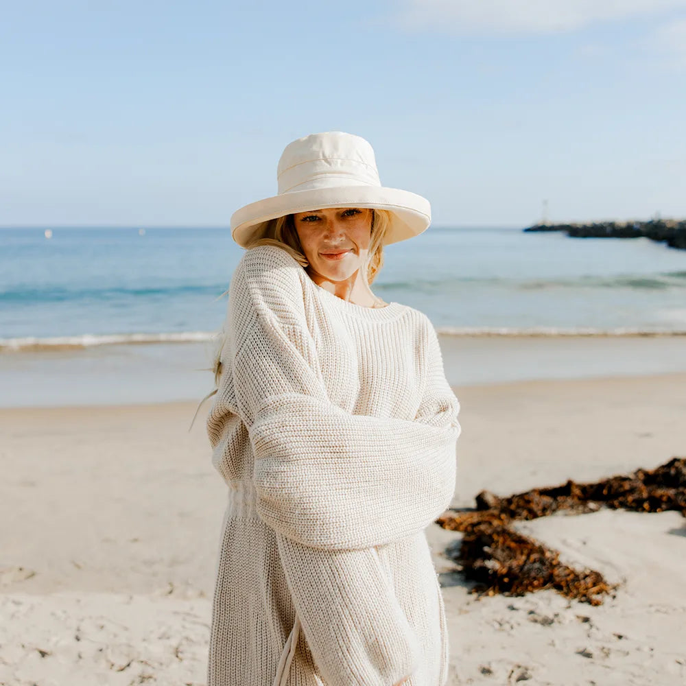 Woman in a long beige cardigan and hat standing on a beach with ocean in the background