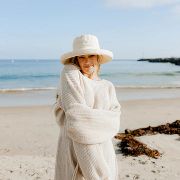 Woman in a long beige cardigan and hat standing on a beach with ocean in the background