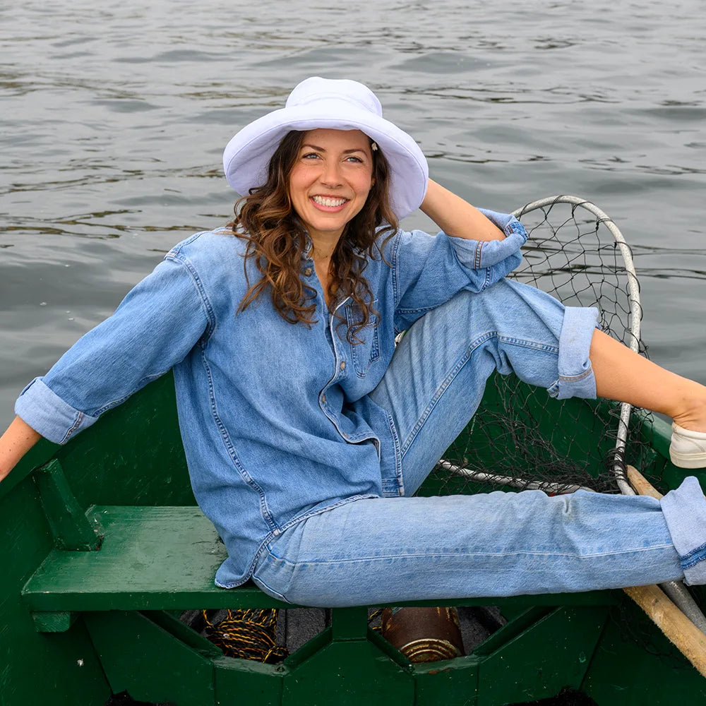 Woman sitting in a green rowboat on water wearing a blue denim outfit and white sun hat.