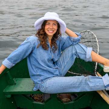 Woman sitting in a green rowboat on water wearing a blue denim outfit and white sun hat.