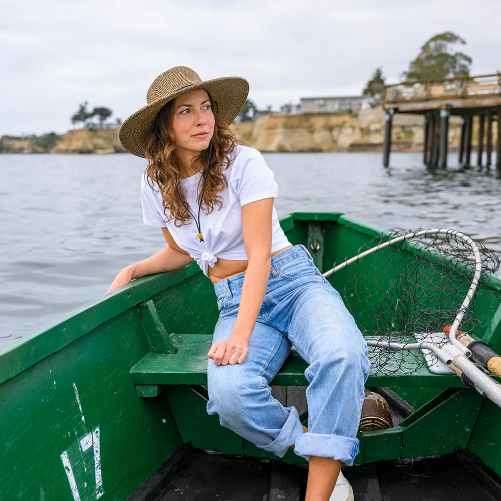 Woman sitting in a green boat on a body of water wearing a straw hat.